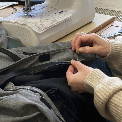 Ein Foto von 2 Frauen-Händen. Die Hände arbeiten mit Nadel und Faden. Die Fau näht eine Jacke. Die Jacke ist grau.
