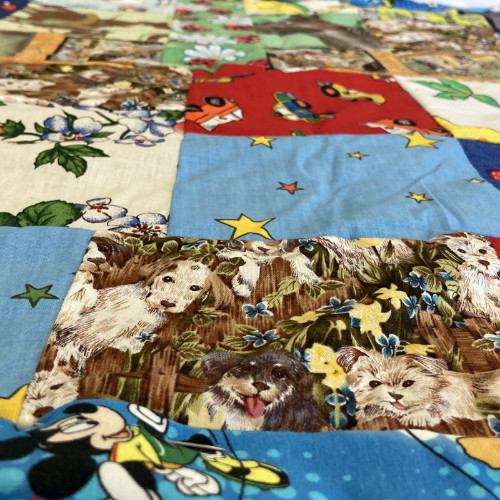 Ein Foto von einer Stoff-Decke. Einzelne Flicken sind auf die Decke genäht. Hunde, Blumen und Comics sind auf den einzelnen Flicken. Die Flicken sind bunt.