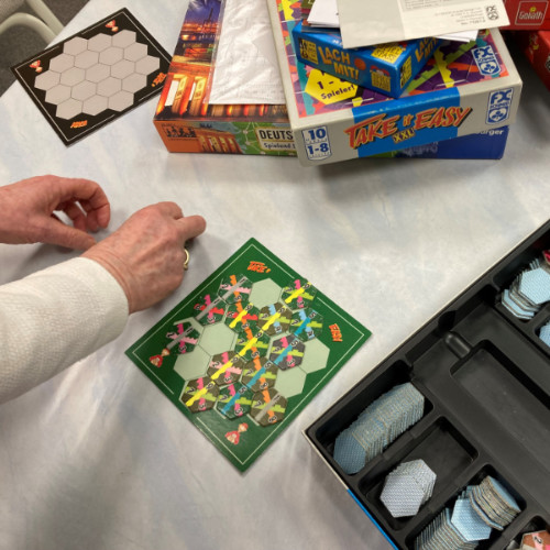 Ein Foto von einem Brett-Spiel auf einem Tisch. Eine Hand legt Figuren auf das Spiel. Weitere Spiele liegen im Hinter-Grund.