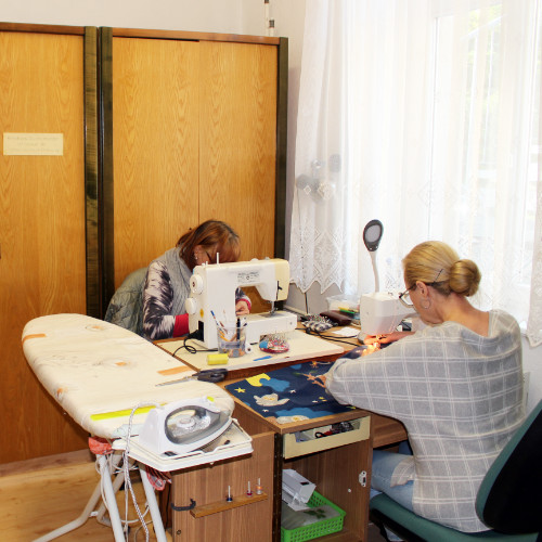 Ein Foto von 2 Frauen an einem Tisch. Die Frauen sitzen an Nähmaschinen. Ein Bügel-Brett steht neben dem Tisch. Ein brauner Schrank steht im Hintergrund.